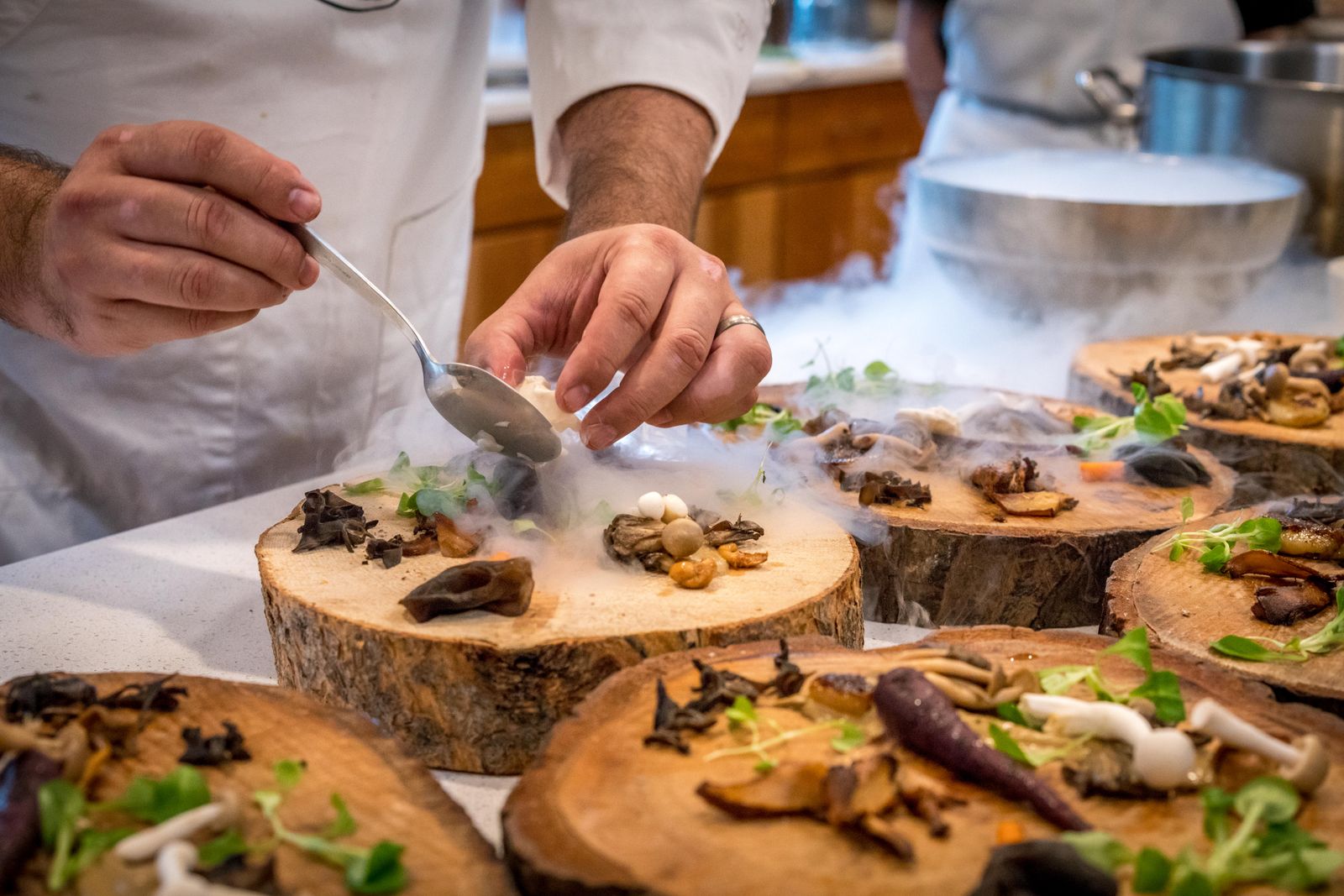 A chef leaning over the counter, spooning smoked cream onto plated wood boards as dry ice smoke drifts across